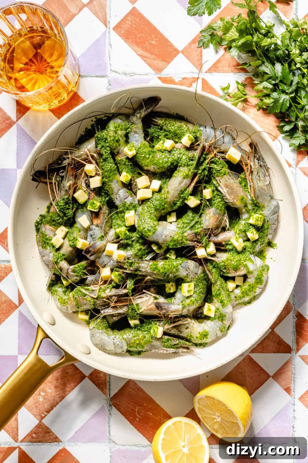 a white skillet with raw shrimp, parsley oil, and cubes of butter with a halved lemon on the side