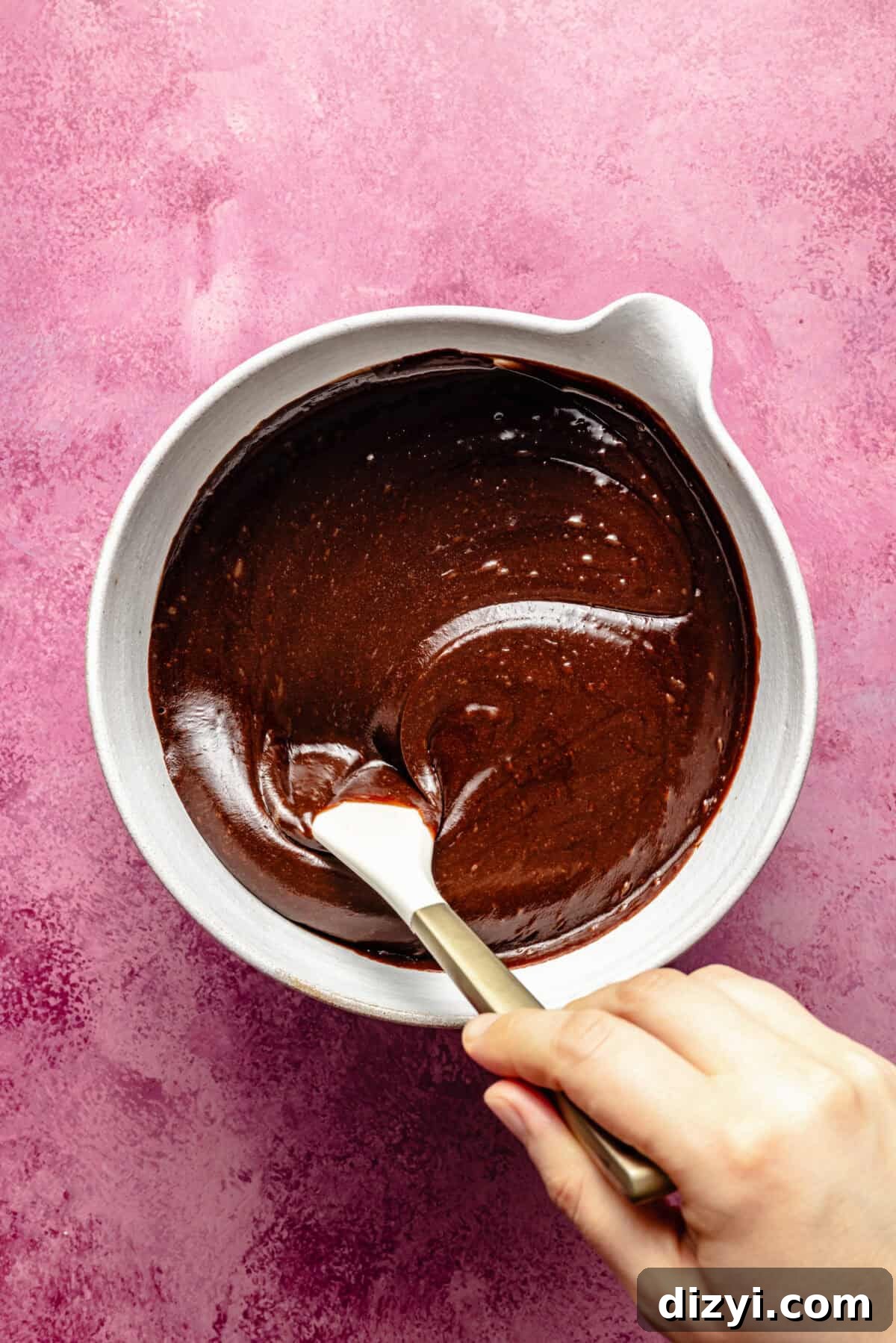 Kiki's Texas Chocolate Sheet Cake Recipe 4 a white bowl and spatula with creamy chocolate frosting on a mauve background