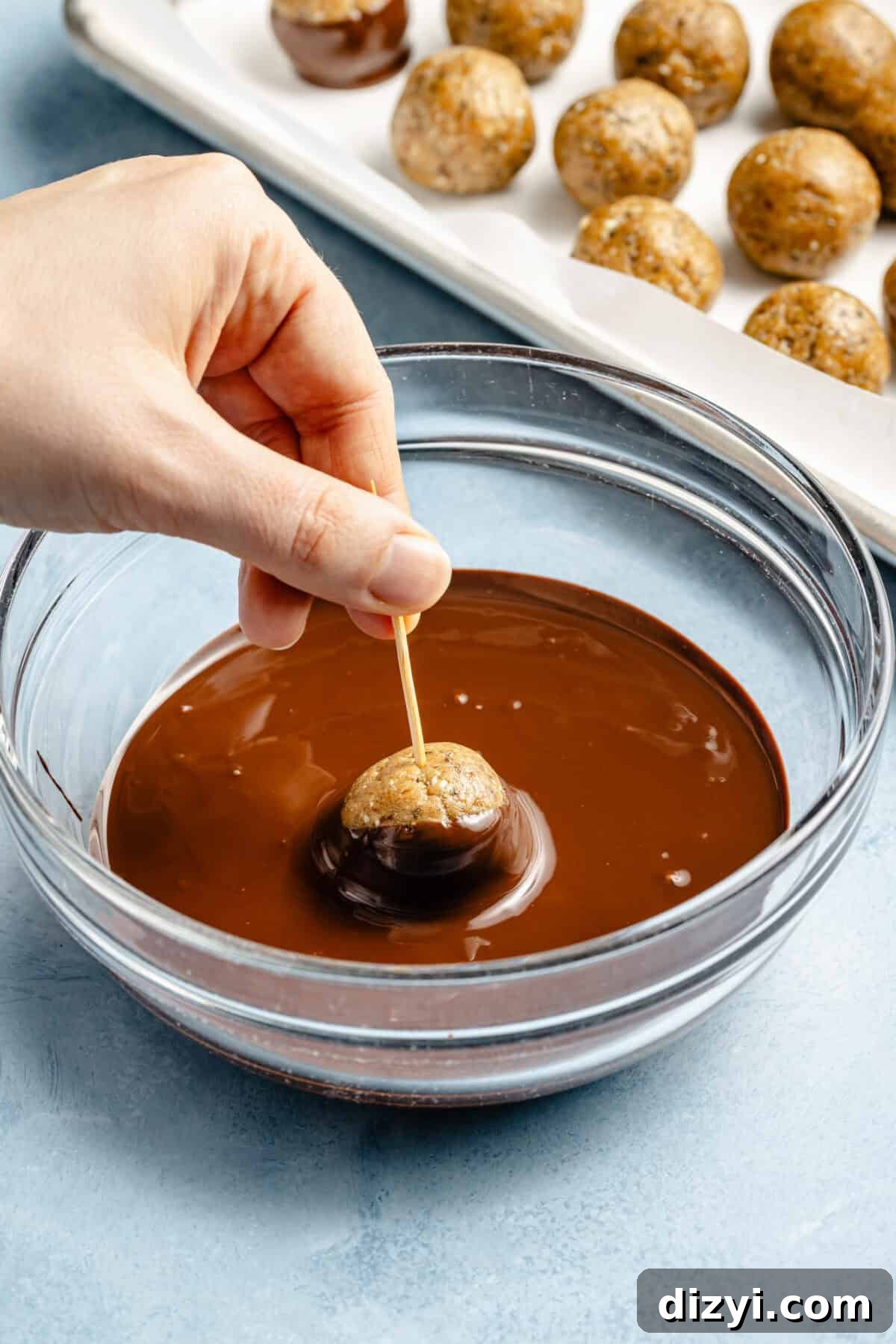 Protein-Packed, Fiber-Rich Buckeye Balls 5 someone dipping a protein ball in melted chocolate in a glass bowl on a light blue background