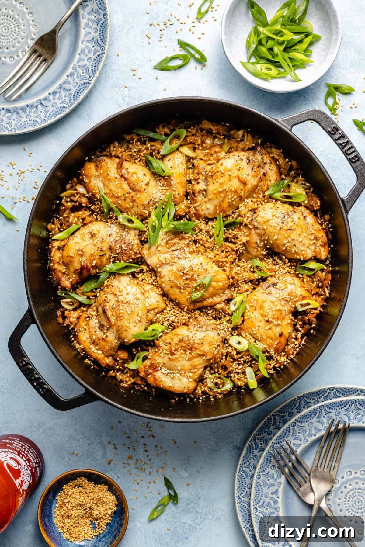 a cast-iron skillet with sesame chicken thighs, rice, and scallions on a light blue background
