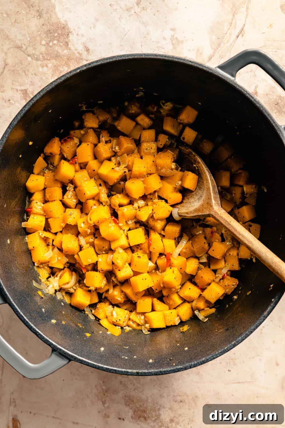 Butternut squash and other ingredients in a black stockpot with a wooden spoon on a countertop.