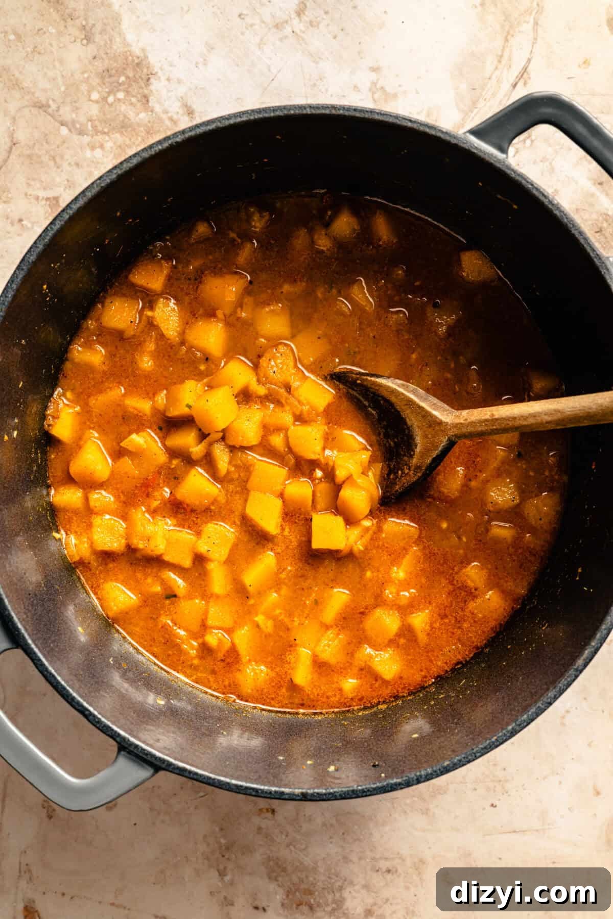 The butternut squash and other ingredients simmering in a black stockpot with a wooden spoon.