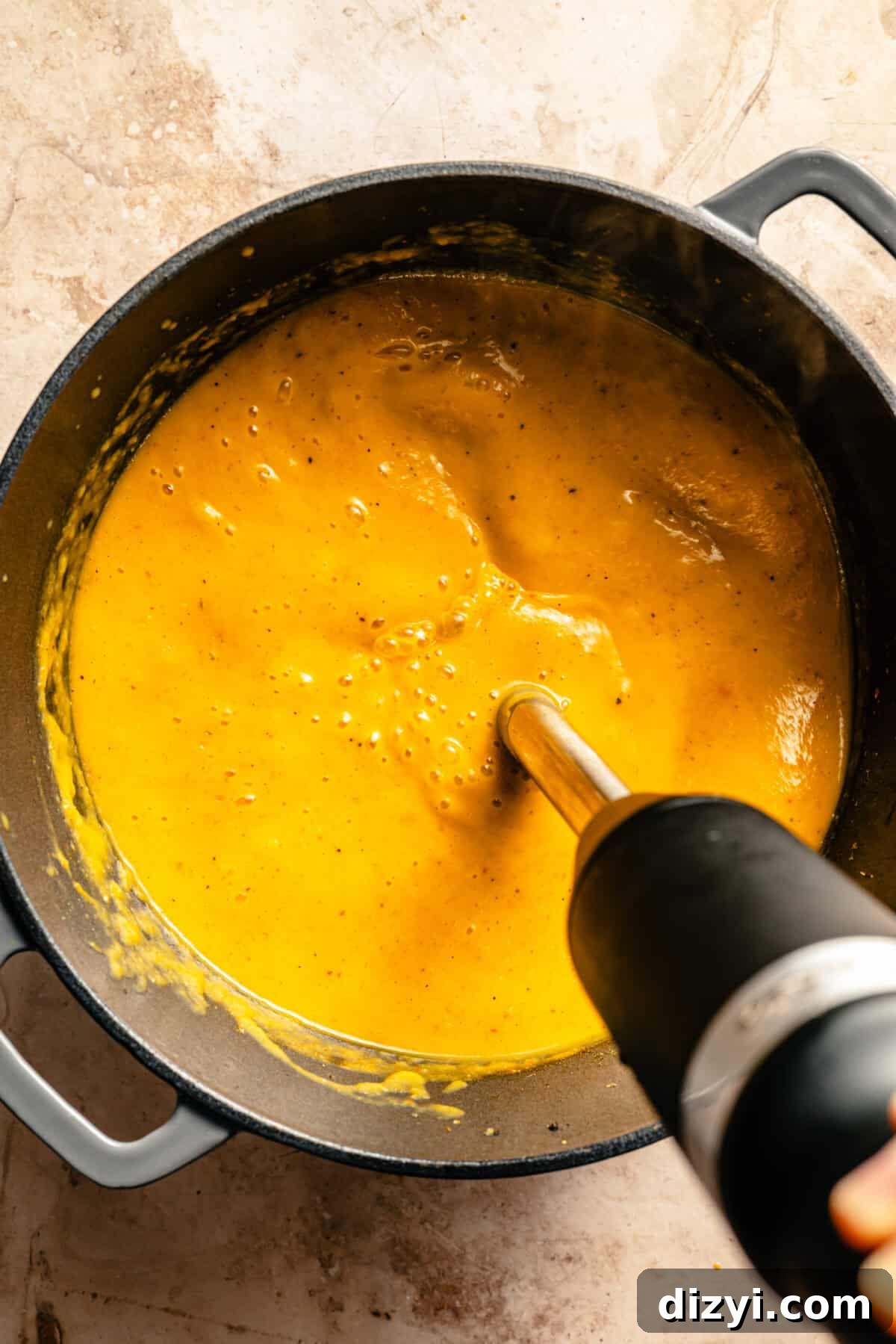 An immersion blender blending the butternut squash ingredients in a black stockpot on a tan countertop.