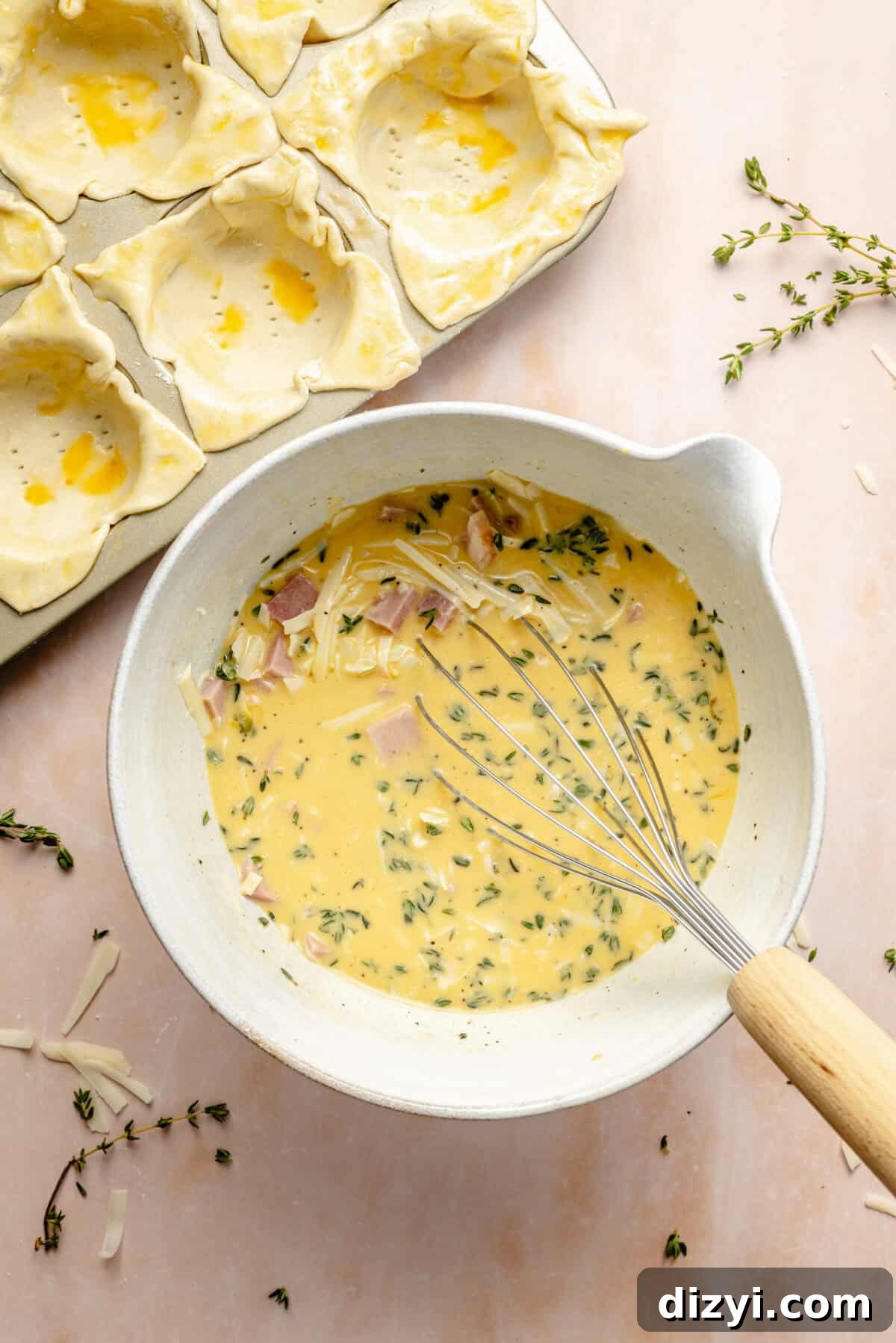 a large white mixing bowl with beaten eggs, fresh herbs, ham, and cheese with a wire whisk