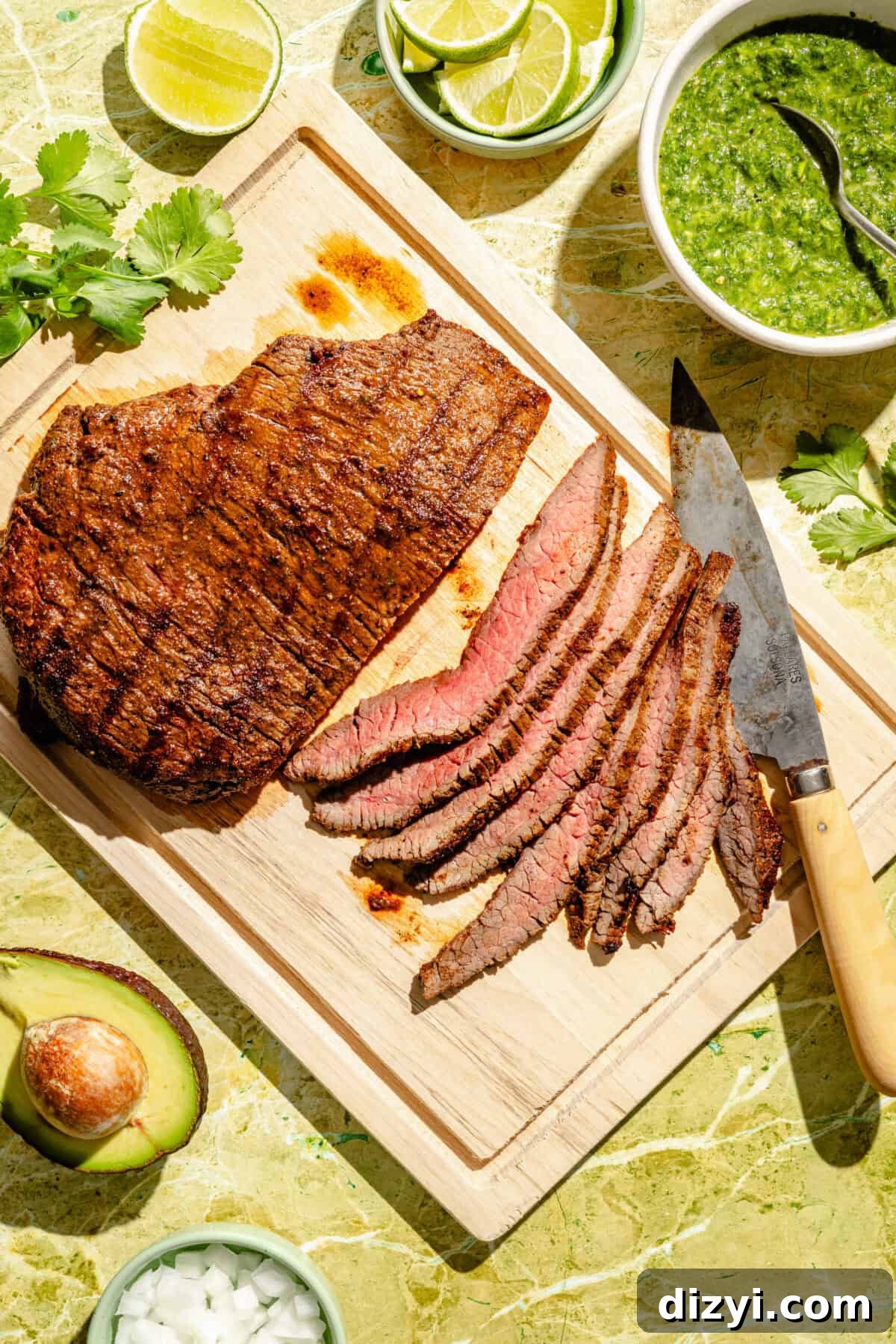 Sliced steak on wooded cutting board with knife. Other ingredients surrounding.