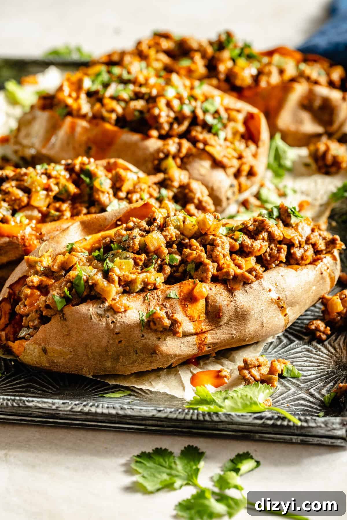 Sloppy Janes: Homemade Sloppy Joe‑Style Sandwiches 4 A close-up shot of a silver patterned baking dish with parchment paper and four finished Sloppy Janes served on top. There is a blue napkin in the top right and sprigs of cilantro around the dish.