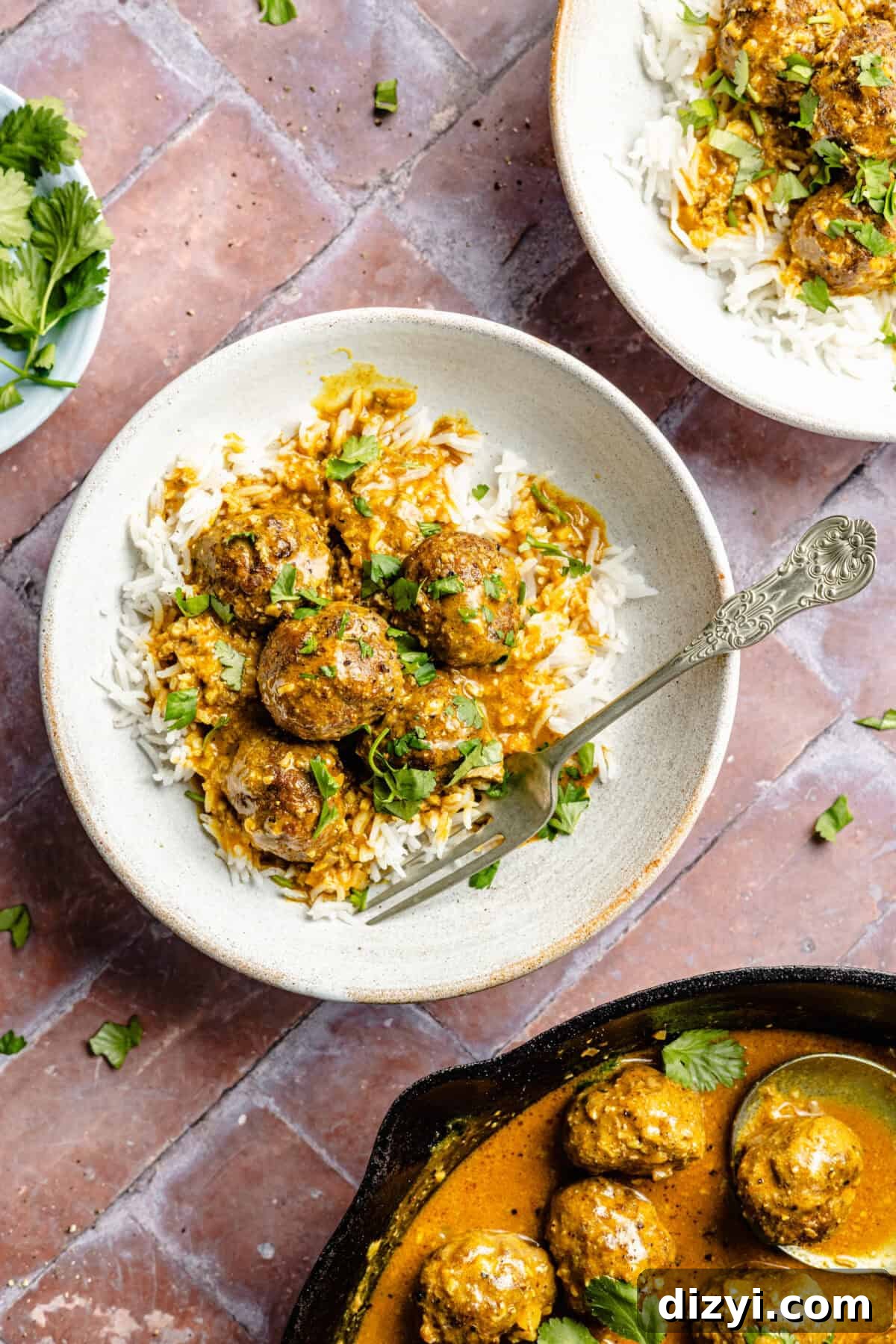 Two white bowls with rice, the finished Easy Tikka Masala Meatballs, and topped with cilantro. The cast iron skillet with the meatballs is visible in the bottom right corner.