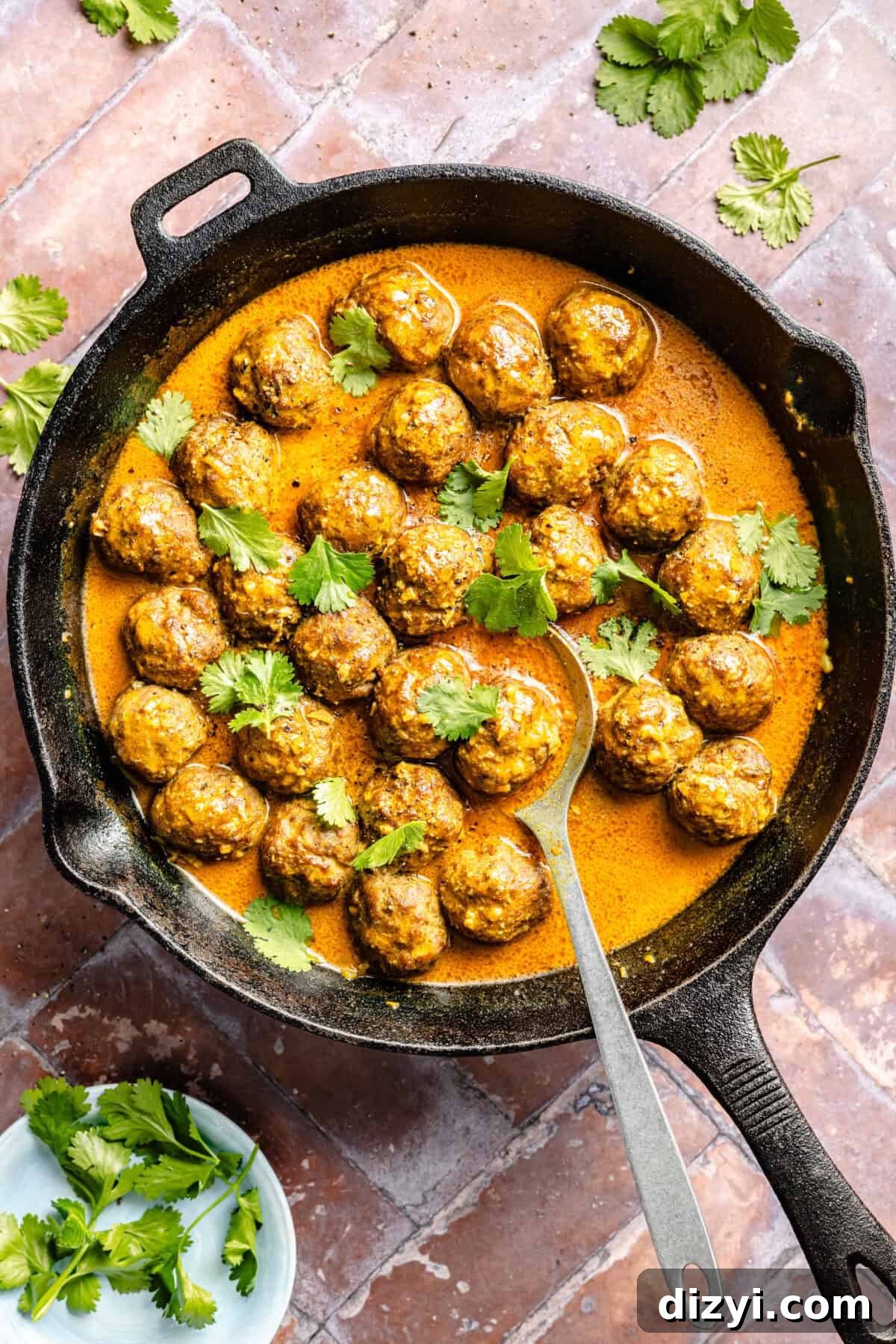 A top-down shot of the cast iron skillet with the saucy Easy Tikka Masala Meatballs, a silver serving spoon, and cilantro topping it. A small white dish of cilantro is in the bottom left corner.