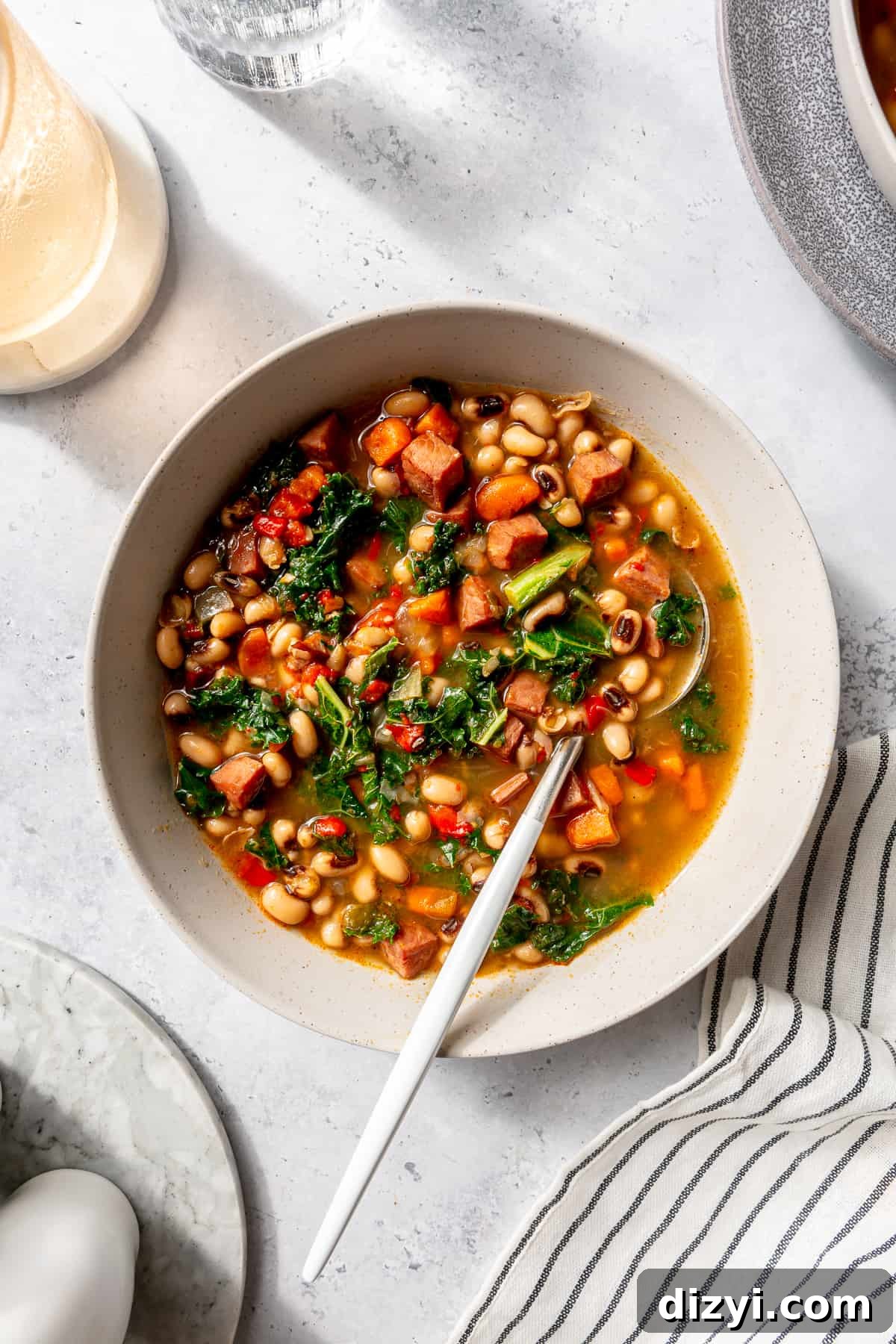 Black-Eyed Pea Soup in a white serving bowl. Spoon resting in bowl. Towel tucked into the bottom corner and plates and cups scattered around.