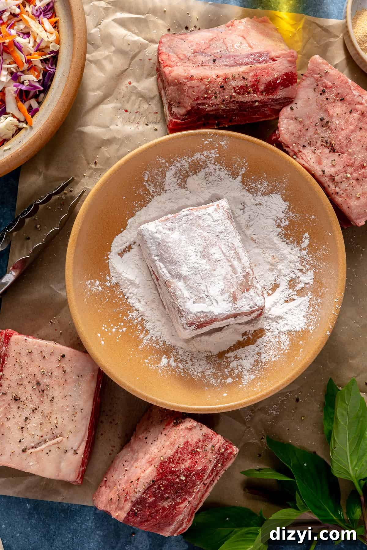 Short rib being dredged in tapioca flour in a yellow bowl. Near the bowl are other seasoned short ribs, a small bowl of slaw, tongs, and parchment paper.